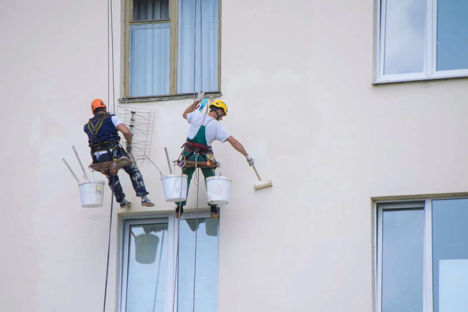 Two exterior painters on harnesses rolling fresh paint onto a cream stucco wall, West Palm Beach, FL