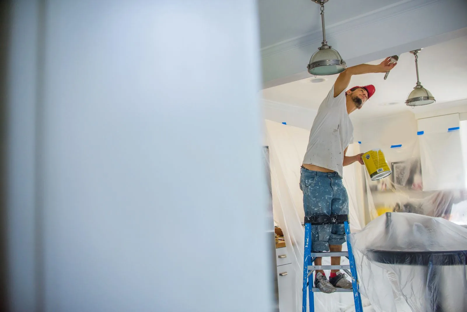 Painter on a ladder cutting in a ceiling line during an interior repaint in West Palm Beach, FL, furniture masked with plastic